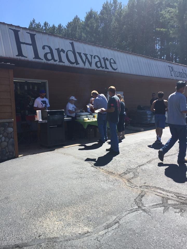The front of Mountain Hardware and Lumber with "Hardware" and "Plumbing" painted along the roofline, customers gathered around a grill and serving tables outside the open door on a summer day.