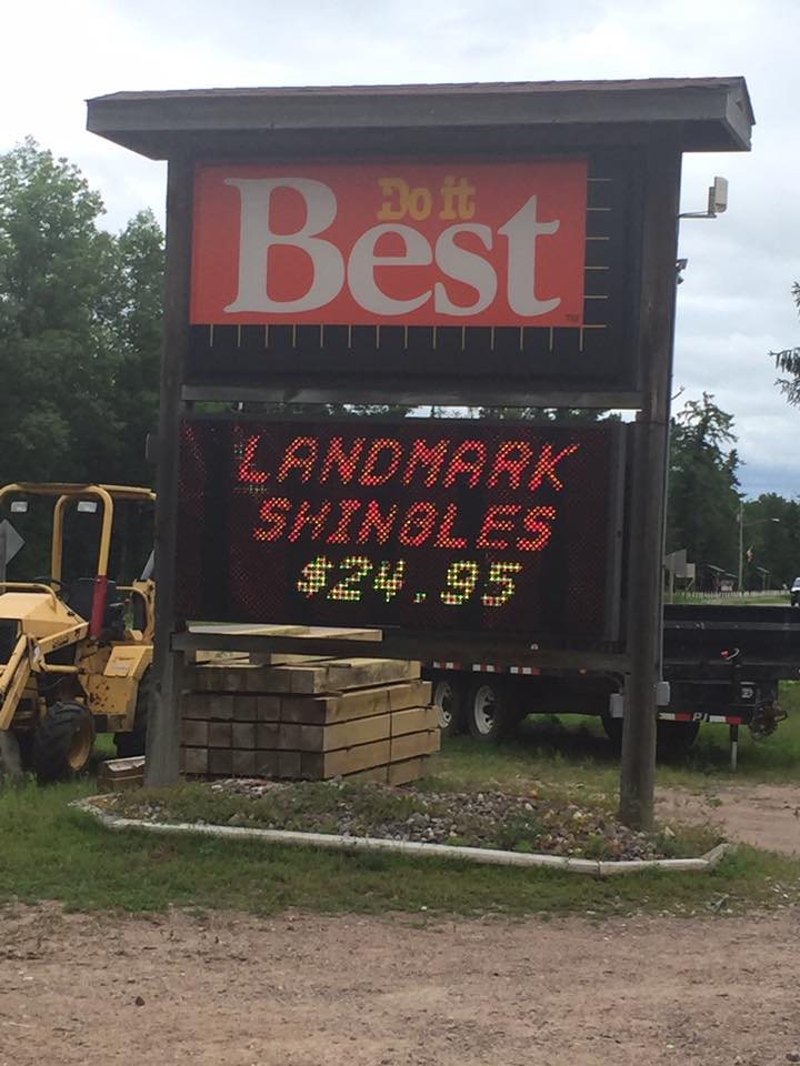The Do It Best roadside sign at Mountain Hardware and Lumber, its LED reader board showing "Landmark Shingles $24.95", with a trencher, stacked timbers and a flatbed trailer in the yard behind.