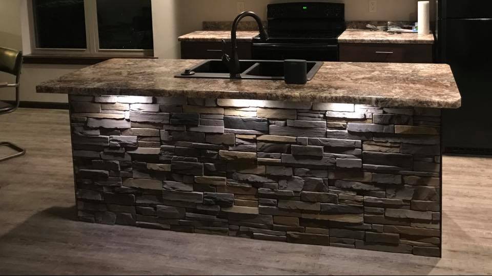 A finished kitchen island faced in stacked stone veneer with a granite top, undercabinet lighting and a black sink, in a newly built kitchen.