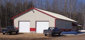 A finished pole barn with white siding and red trim, two overhead doors, and a pair of pickup trucks parked on the gravel outside.