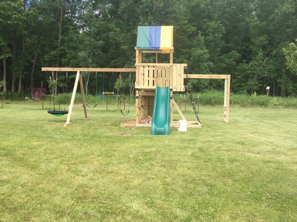 A wooden swing set and playhouse with a green slide and a canopy, built from dimensional lumber, standing on mown grass at the edge of the woods.