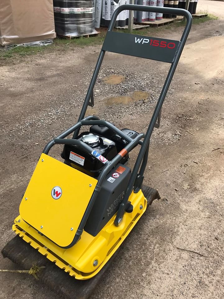 A yellow Wacker Neuson WP1550 compactor plate standing on gravel in the yard, with pails of roof coating stacked on pallets behind it.