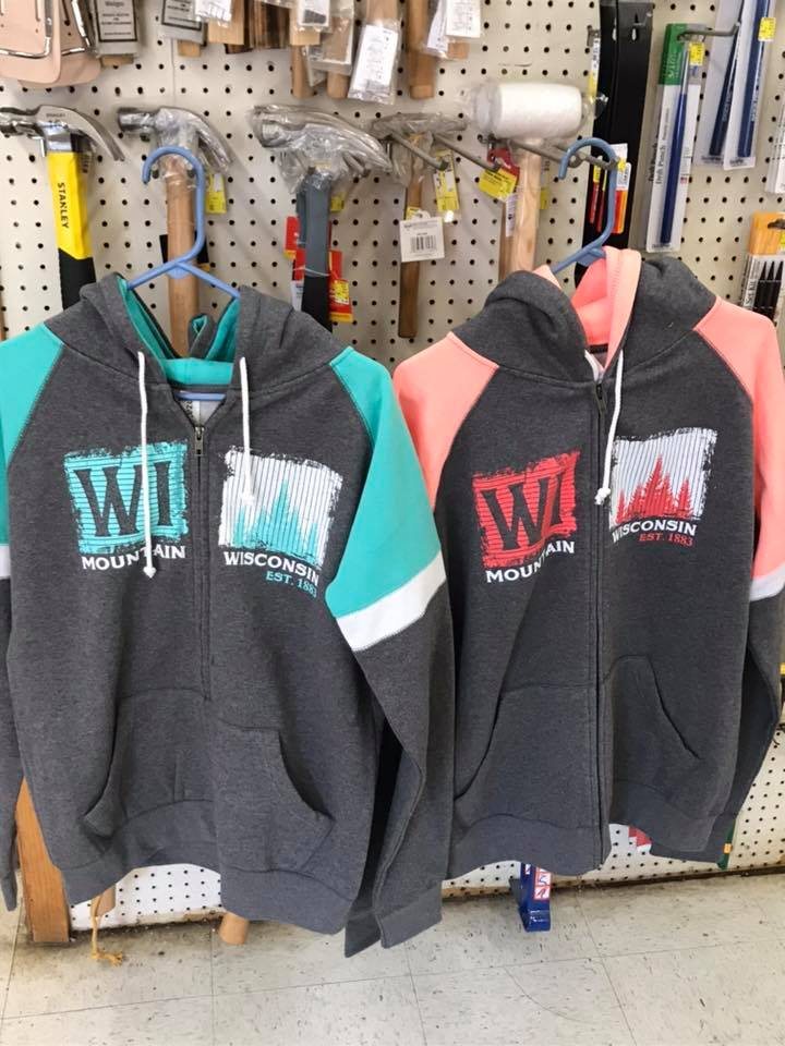 Two Mountain, Wisconsin hooded sweatshirts hanging in front of a pegboard wall of hammers, mallets and hand tools inside the store.